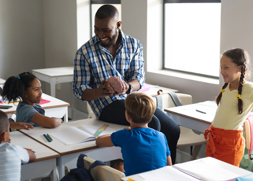 Black, male teacher sits on a desk holding a brain model talking with young students. 